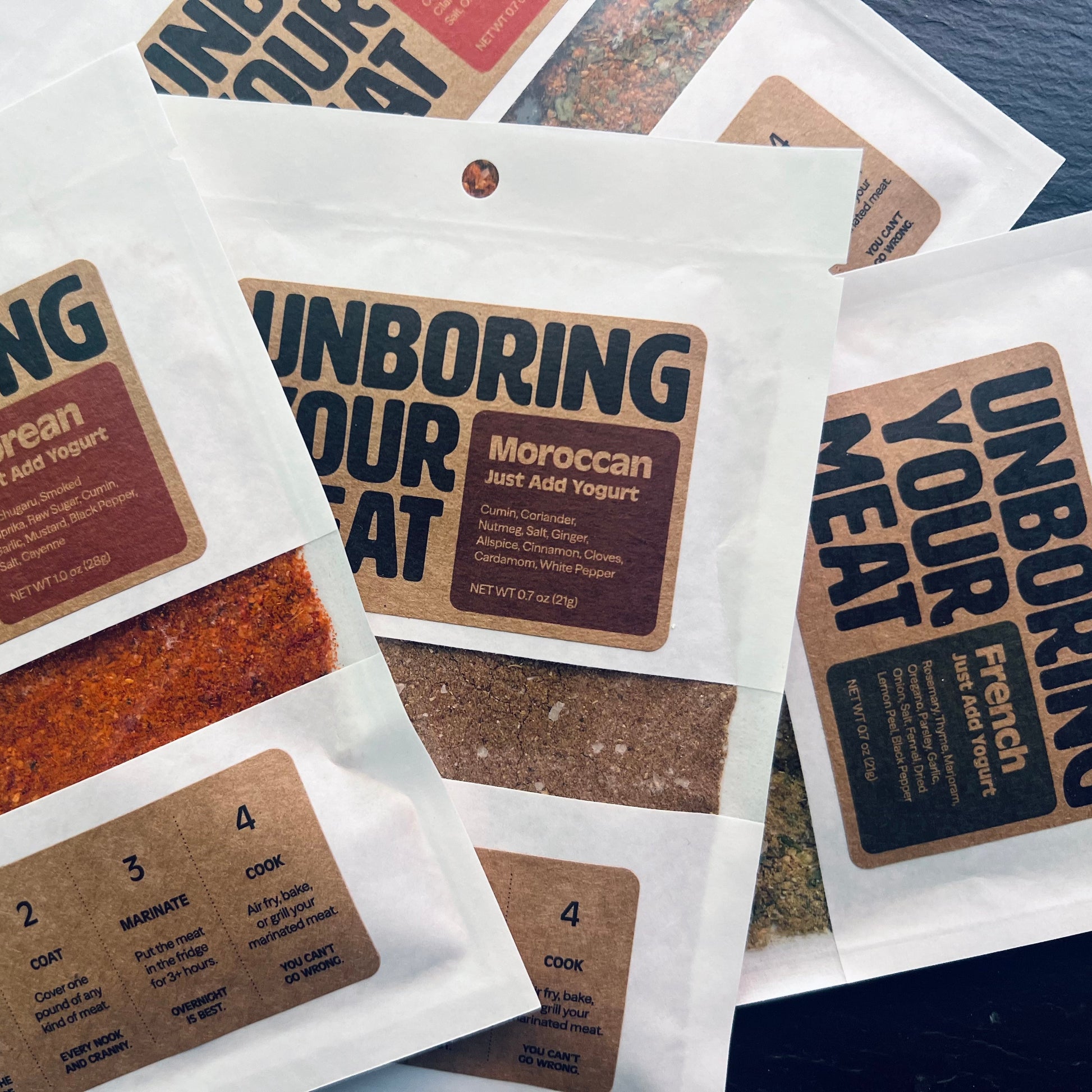 Packets of 'Unboring Your Meat' with different spice blends on a blue surface.